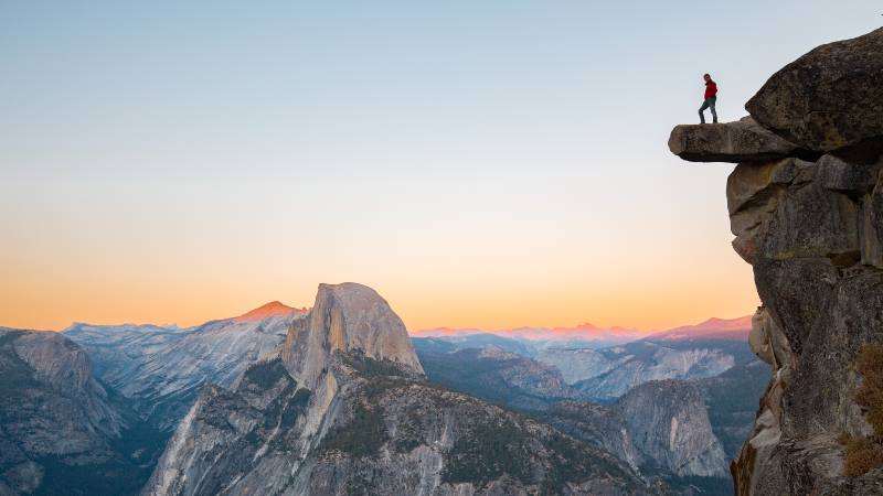 A-fearless-hiker-is-standing-on-an-overhanging-rock-enjoying-the-view-towards-famous-Half-Dome-at-Glacier-Point-overlook-in-beautiful-evening-twilight-Yosemite-National-Park-California-USA